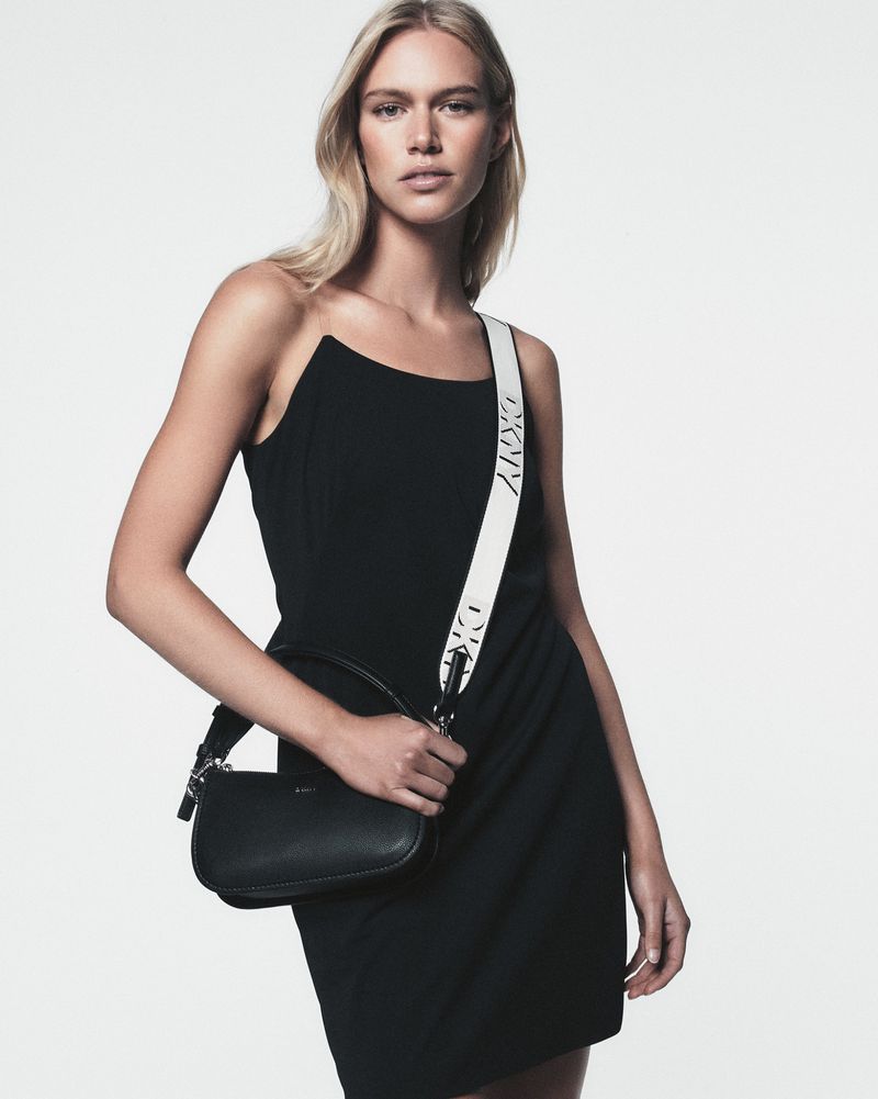 Fashion shot of a blonde woman wearing a black dress, holding a small black purse with a white strap against a plain white backdrop.