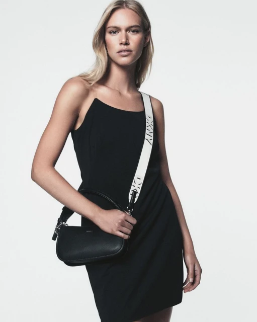 Fashion shot of a blonde woman wearing a black dress, holding a small black purse with a white strap against a plain white backdrop.