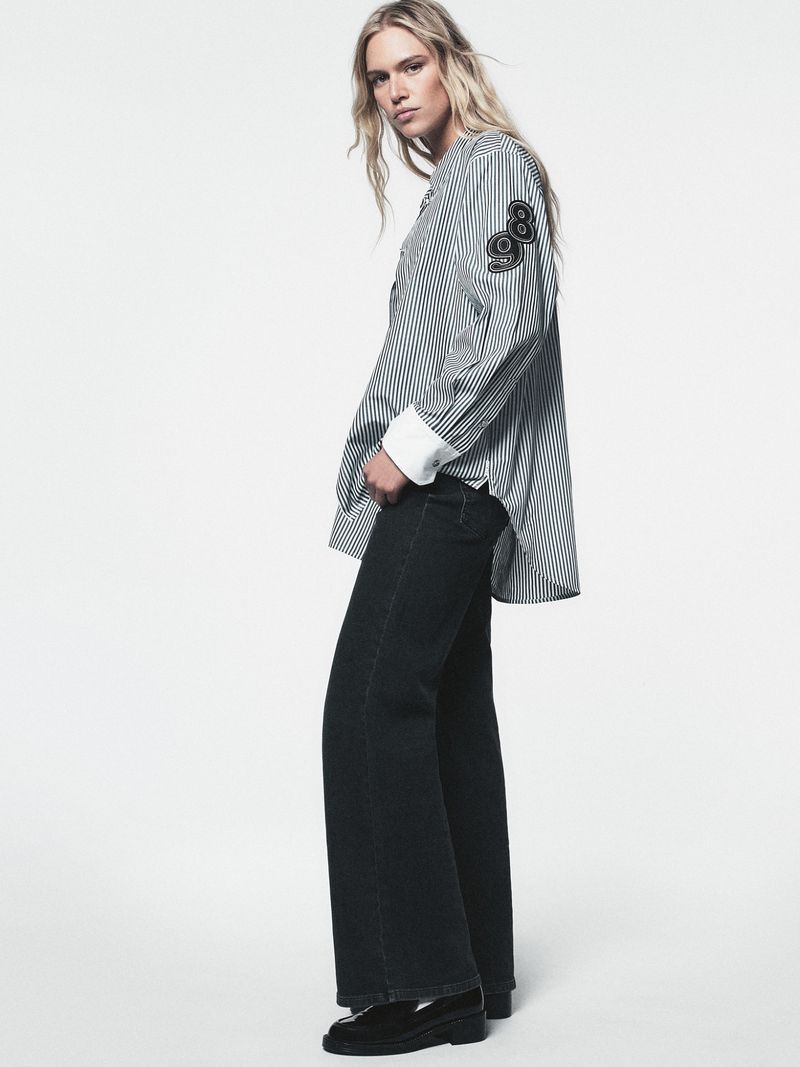 Fashion model in a striped shirt and wide-leg jeans posing in a photo studio. Full-length shot of a woman in an outfit against a plain background.