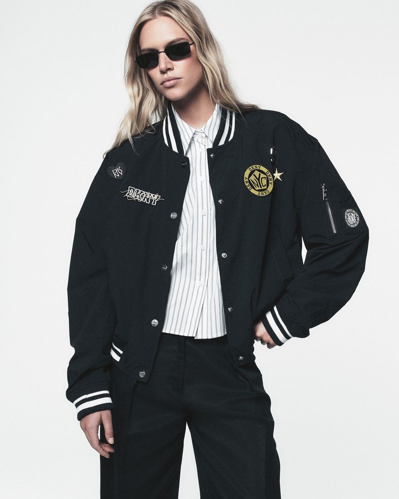 Woman in a black and white button-down shirt and black bomber jacket with patches poses in a studio with a white background.