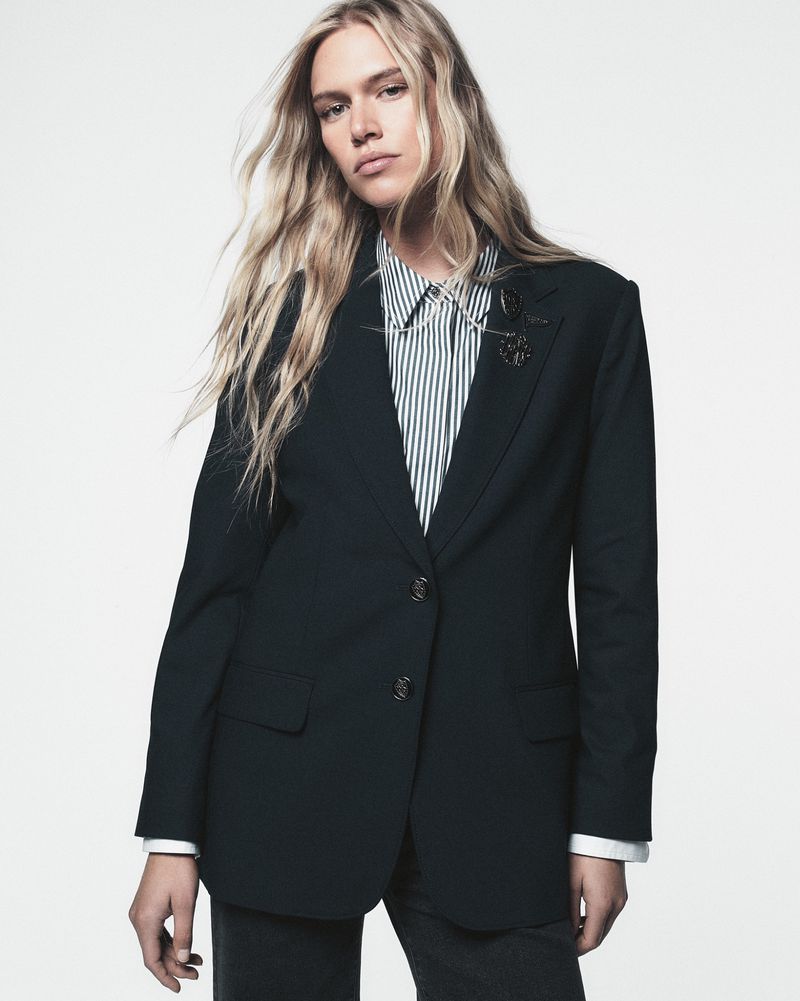 Studio portrait of a blond female model in a dark green blazer with a striped button-down shirt. Fashion shot with a plain white background.
