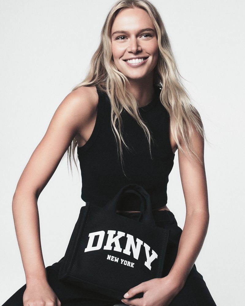 Smiling blonde model poses with a black DKNY New York tote bag, wearing a black tank top and dark pants against a white backdrop.