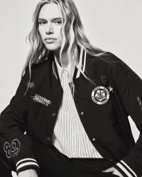 Model wearing a black bomber jacket over a pinstripe shirt against a neutral grey background for DKNY SP26 campaign