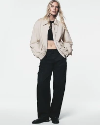 Nikki McGuire wearing a beige zip jacket, black crop top, and wide-leg black trousers on a white studio background.