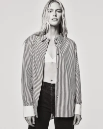 Nikki Mcguire wearing an open striped button-down shirt over a white bralette and dark jeans against a studio backdrop.