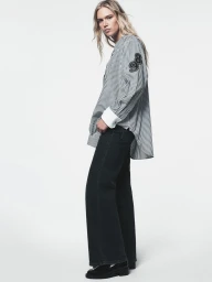 Model Nikki Mcguire wearing a striped oversized button-down shirt and wide-leg black jeans against a bright white background.