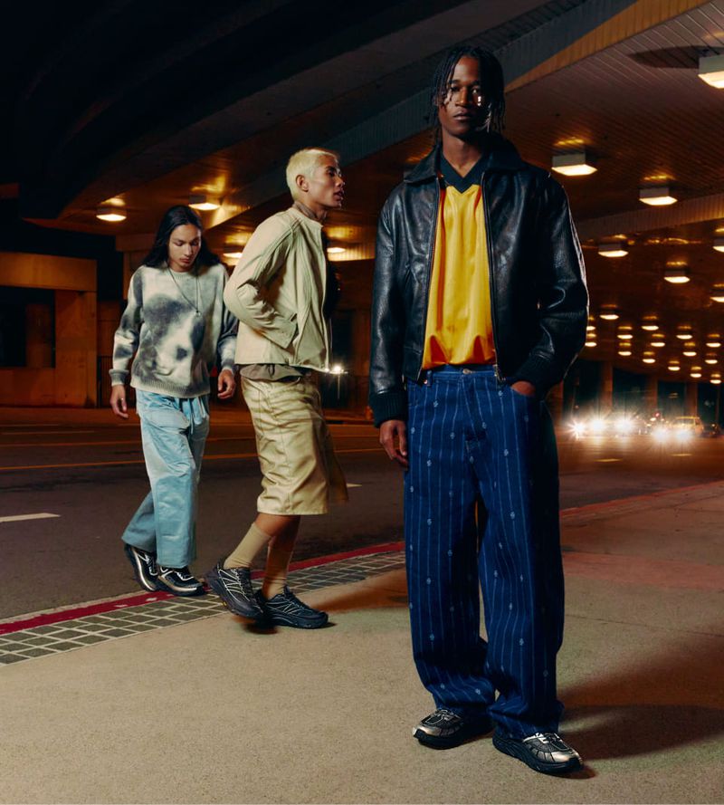 Three adult male models pose at night on a sidewalk under an overpass, showcasing street style with leather jackets and blue trousers.