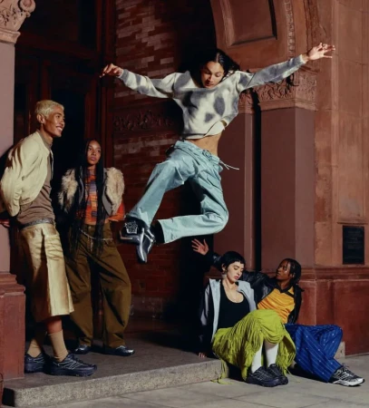 Group of six young adult diverse models in casual clothing posed in an urban setting, with one leaping in the air by brick building.