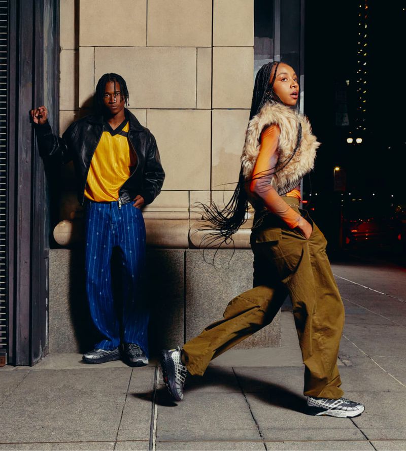 Two adults in stylish clothing stand in an urban environment at night, with architectural details and muted, neutral color tones.