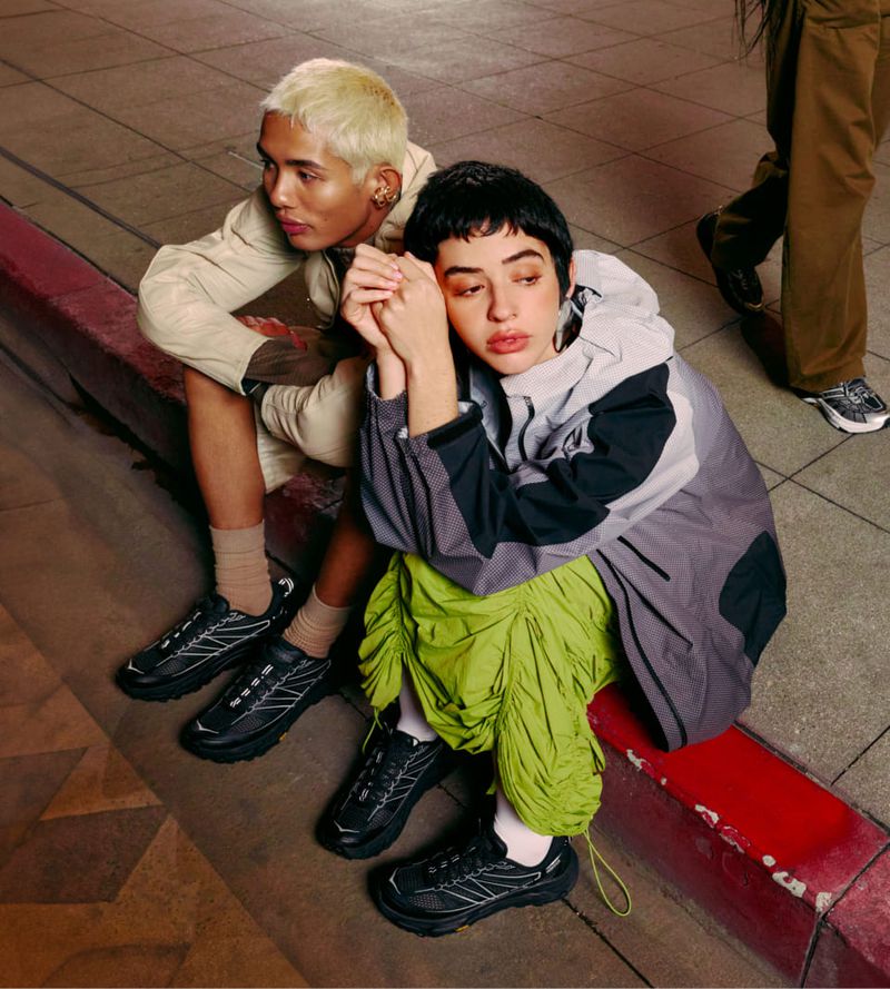 Two people in athletic wear sitting on a curb next to black sneakers in an urban setting at night, with a third person walking by.