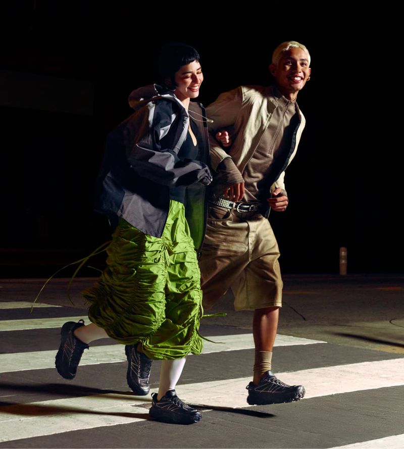 Two athletic adults, a woman in green and a man in beige, run on a pedestrian crossing in athletic shoes, captured in a medium shot.