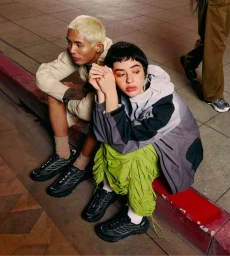 Two models seated on a concrete sidewalk curb wearing black technical running sneakers and modern streetwear.