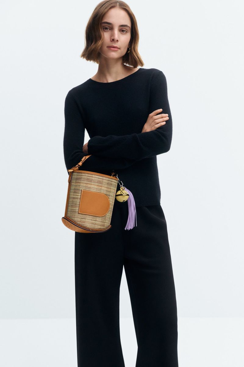 Studio shot of a brunette woman in a black sweater and pants, posing with a woven bucket bag against a white backdrop.