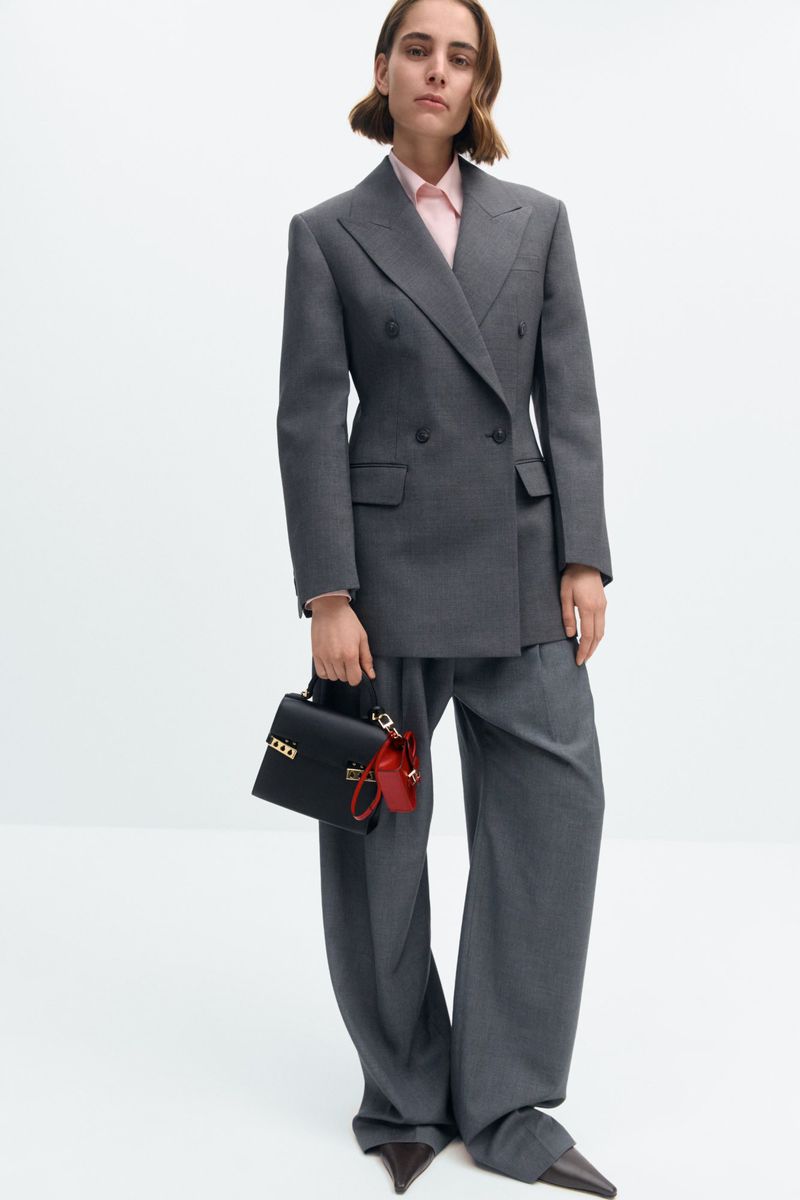 Adult woman posing in a grey pantsuit with a black top-handle bag and red charm in a brightly lit photo studio with a white backdrop.