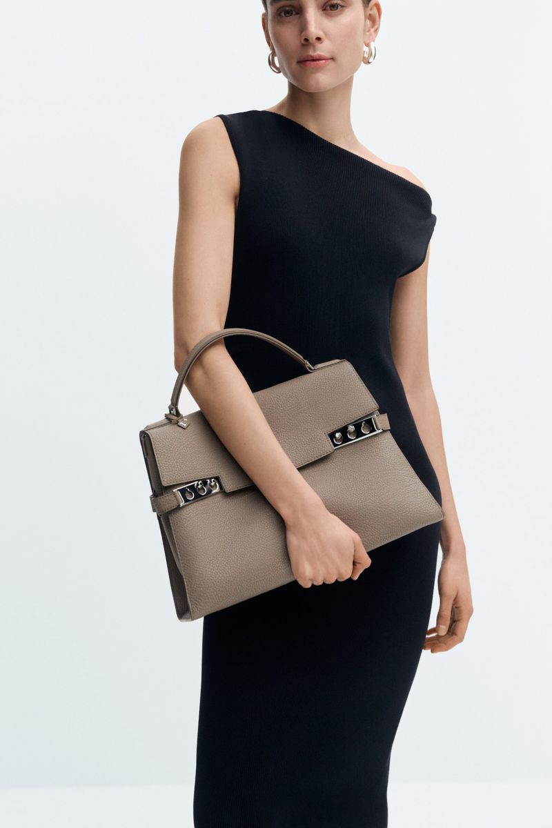 Model Romy Schonberger wearing a black asymmetrical dress while holding a taupe Delvaux leather handbag against a white background.
