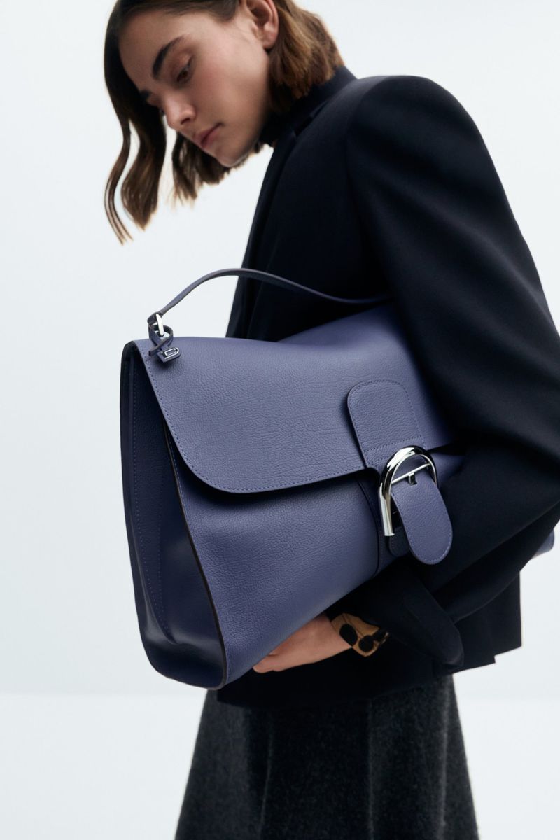 Adult female wearing a black blazer holding a blue leather satchel bag with silver hardware in a minimalist studio setting.