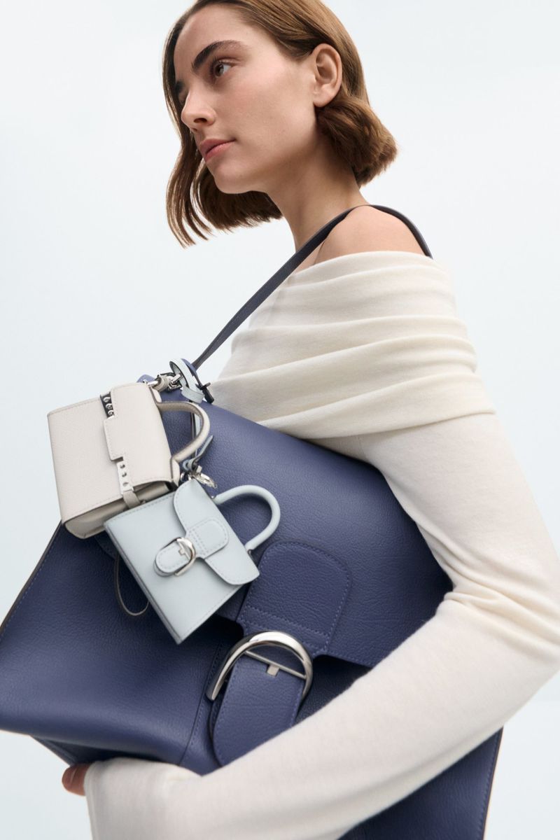 Attractive woman in a beige sweater carries a large navy blue leather satchel with attached small coin purses in a bright studio setting.