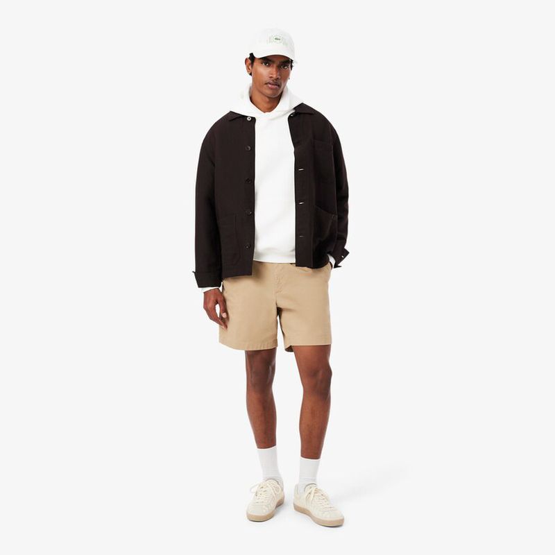 Full shot of a man in a white hoodie, brown shorts, jacket, cap, and sneakers against a white background, showcasing casual fashion.