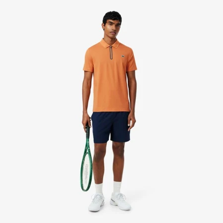 Athletic man in an orange polo shirt and navy shorts, standing on a white background, holding a green tennis racket, looking at the camera.