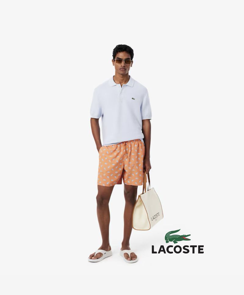 A light-skinned male model stands in a white studio wearing a blue polo shirt, patterned shorts, sunglasses, and sandals, holding a tote bag.