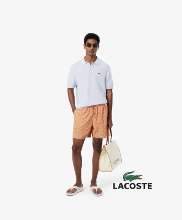 A light-skinned male model stands in a white studio wearing a blue polo shirt, patterned shorts, sunglasses, and sandals, holding a tote bag.