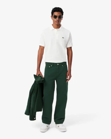 A full-length shot of a man wearing a white polo shirt, green pants, and sunglasses against a white backdrop in a fashion setting.