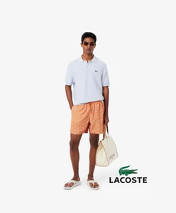 Model Deepak Gupta posing in a pale blue polo shirt, orange patterned shorts, and sandals with a tote bag on a white background.