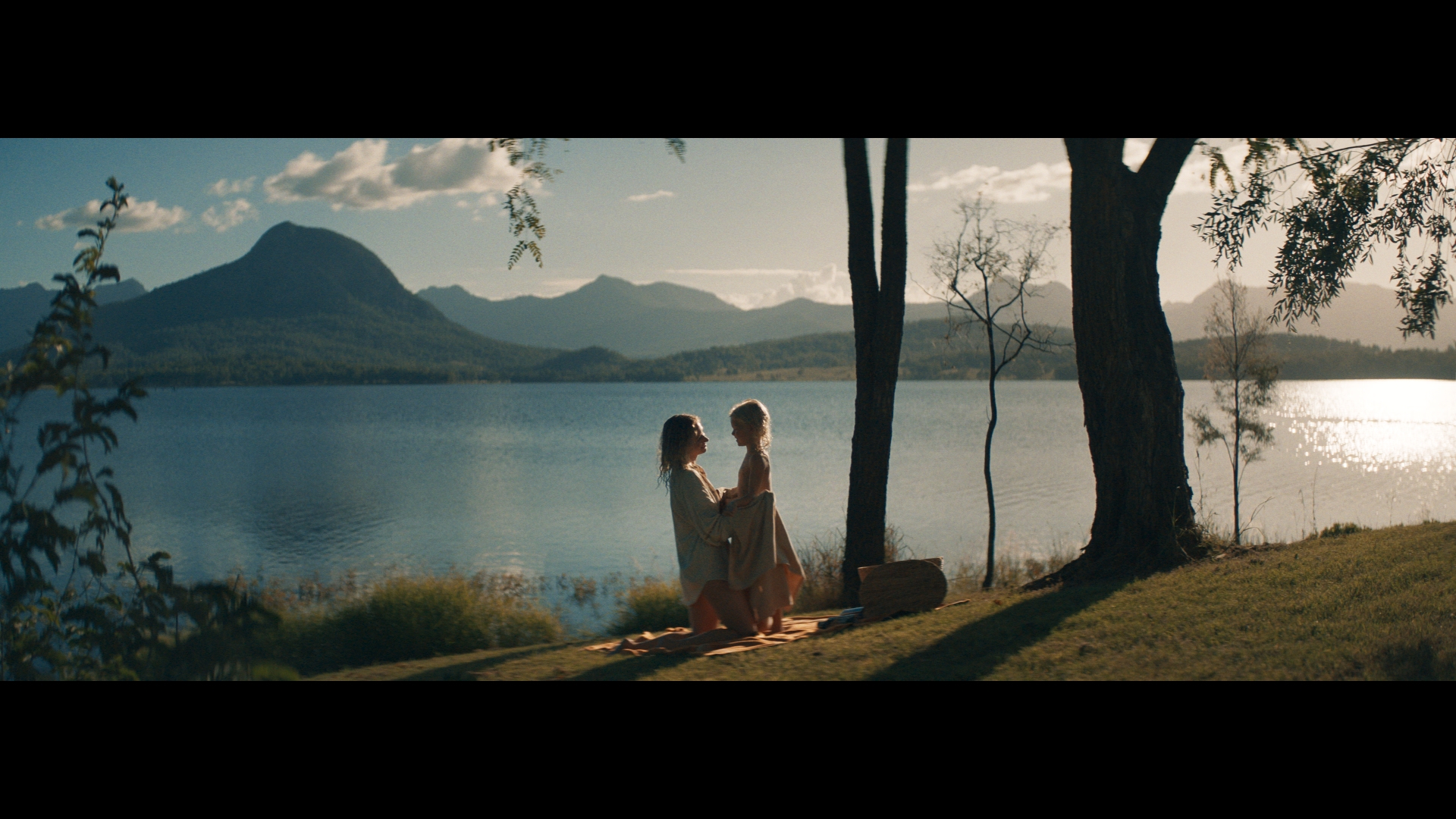Two people standing together on a grassy lakeshore near trees with mountains and water in the background during golden hour.
