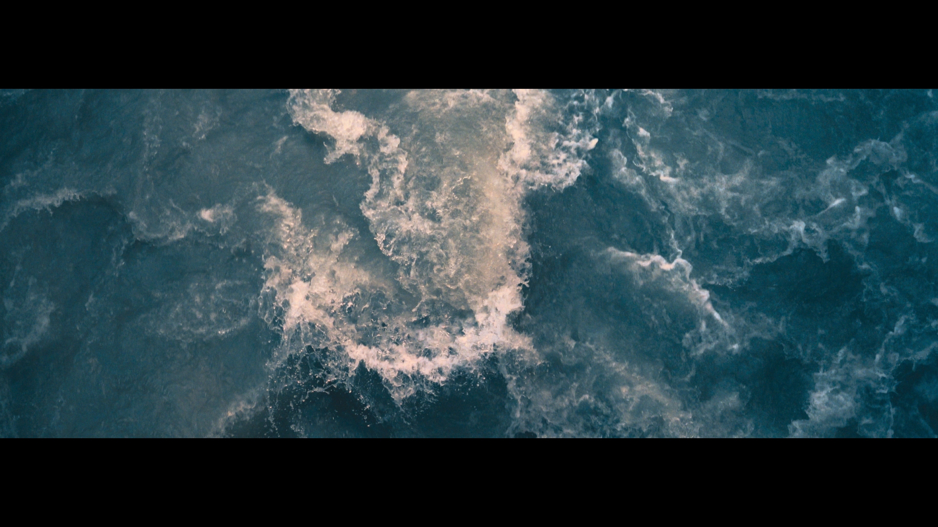 Aerial view of ocean water with a turbulent surface of teal blue and white foam, capturing natural movement and contrasting textures.