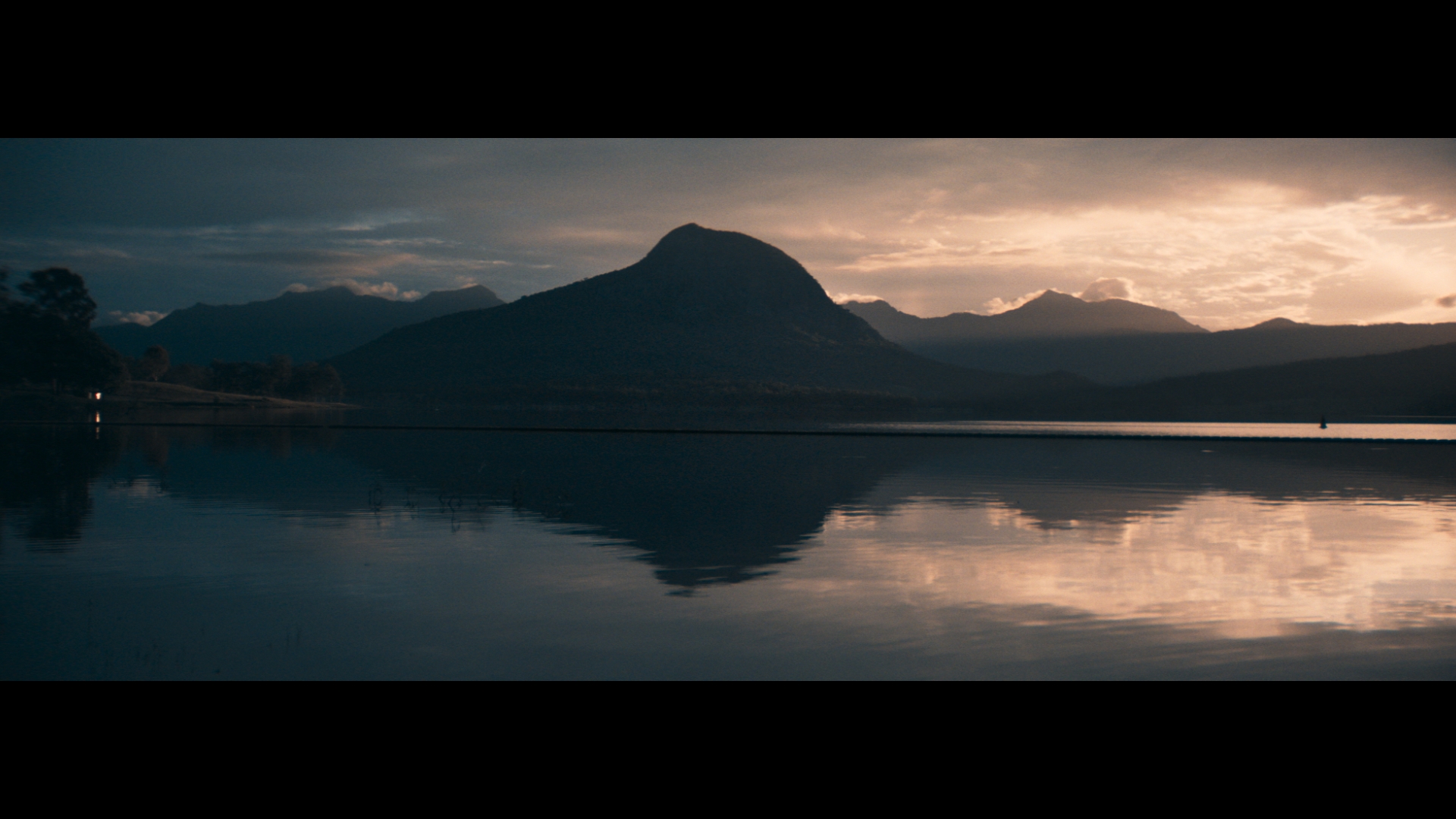 Wide scenic view of calm water reflecting dark mountain silhouettes beneath a dramatic sunset sky with warm golden light.