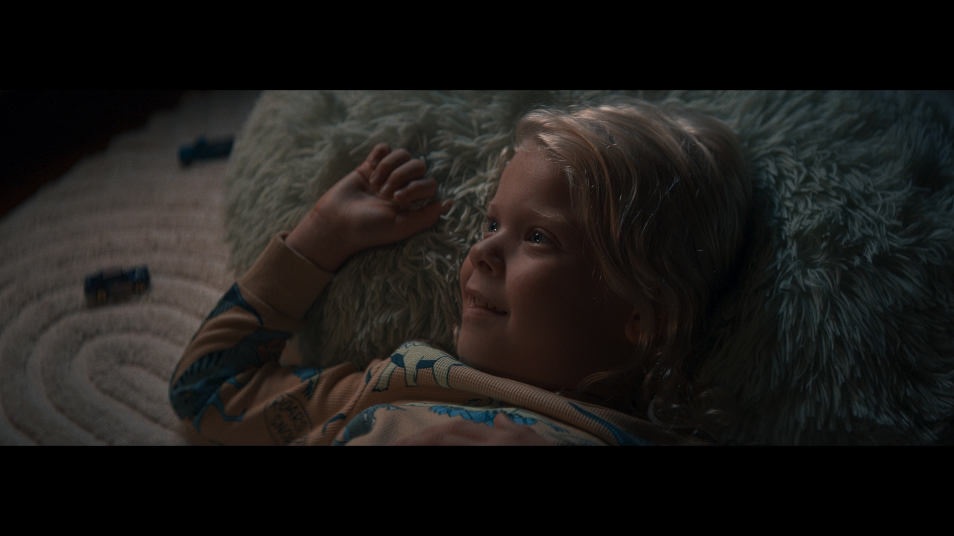 Blonde child lies on a fluffy rug, looking up with a slight smile, wearing patterned pajamas, two toy cars visible in the background.