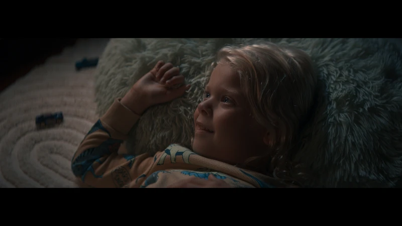Blonde child lies on a fluffy rug, looking up with a slight smile, wearing patterned pajamas, two toy cars visible in the background.