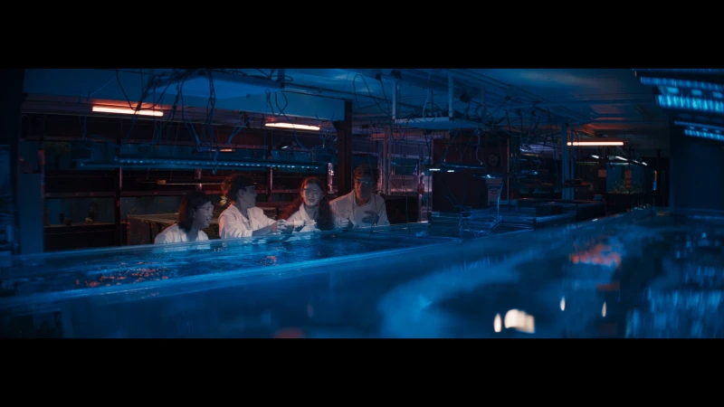 Four people in lab coats are gathered at an illuminated water tank in a dimly lit, high-tech research environment at nighttime indoors.