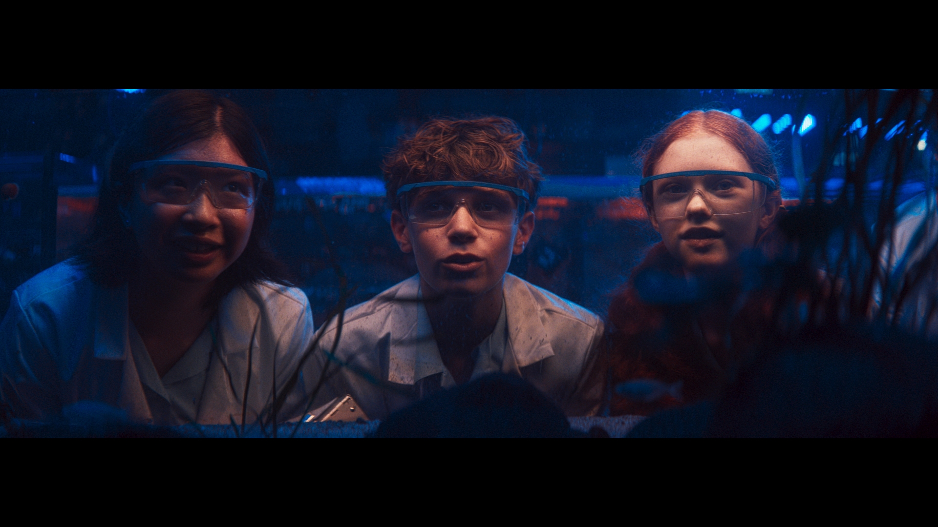 Three children wearing lab coats and safety glasses look at something in front of them in a dimly lit room with blue lighting.