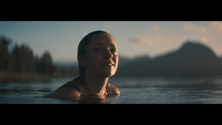 Woman with wet hair smiling while wading in a lake with mountain silhouettes under an evening sky.