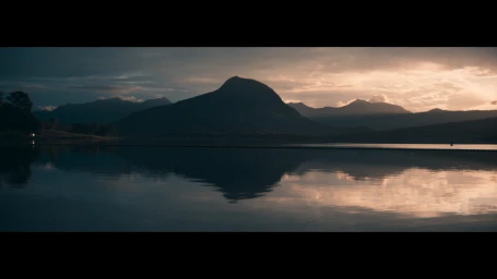 Wide scenic view of calm water reflecting dark mountain silhouettes beneath a dramatic sunset sky with warm golden light.