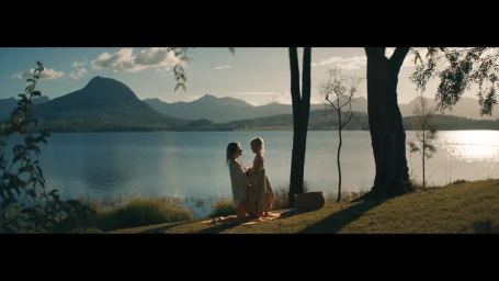 Two people standing together on a grassy lakeshore near trees with mountains and water in the background during golden hour.