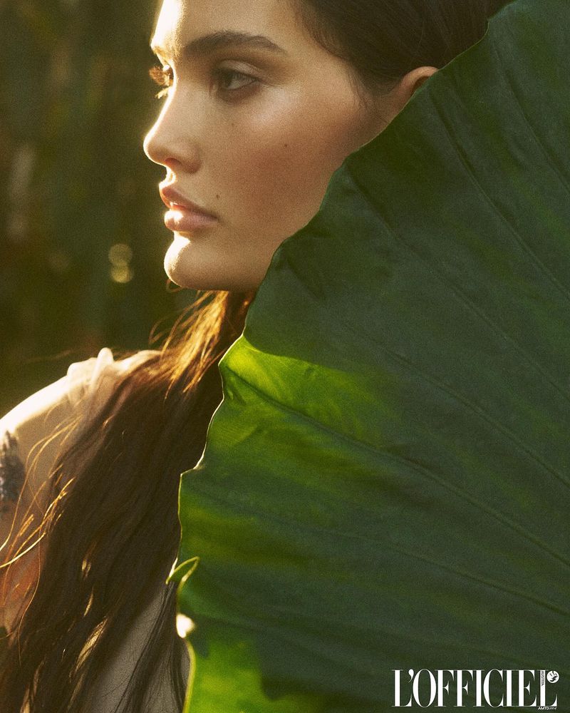 Close-up portrait of a woman's face partially obscured by a large, vibrant green leaf, bathed in golden sunlight.