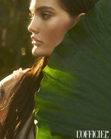 Close-up portrait of a woman's face partially obscured by a large, vibrant green leaf, bathed in golden sunlight.