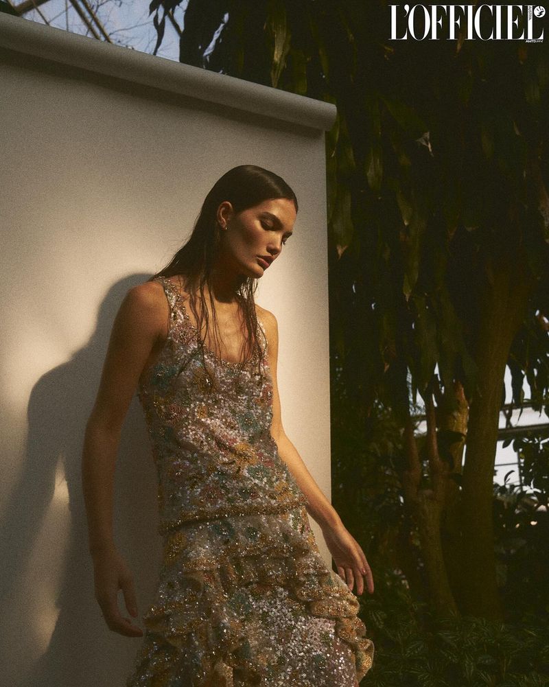 A woman with wet hair poses in a sequined gown in front of a white wall and dark green foliage, bathed in soft lighting.