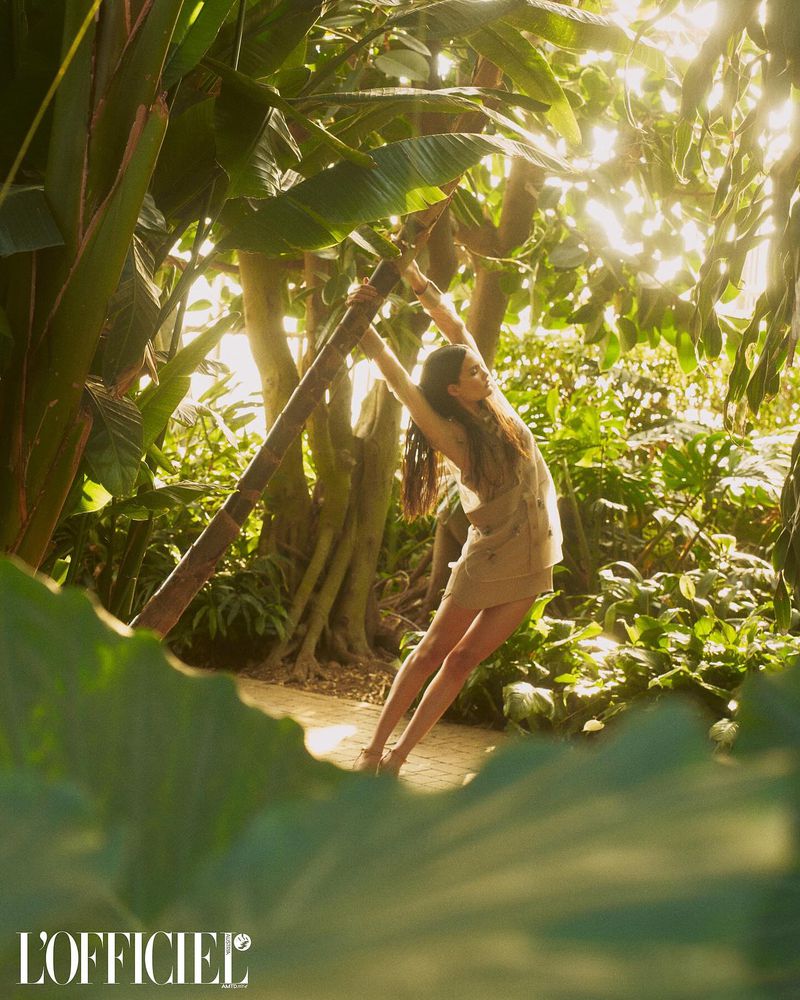 A woman poses in a beige dress, surrounded by lush greenery and large leaves, bathed in soft, natural light in an outdoor setting.