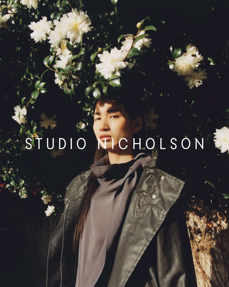 Portrait of Asian woman in oversized grey coat standing outside with white flowers and dark foliage in the background in soft, diffused lighting.
