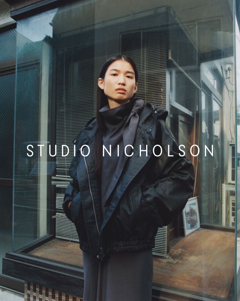 A woman with long black hair poses in front of a reflective window wearing a dark jacket and grey dress for a fashion shoot.