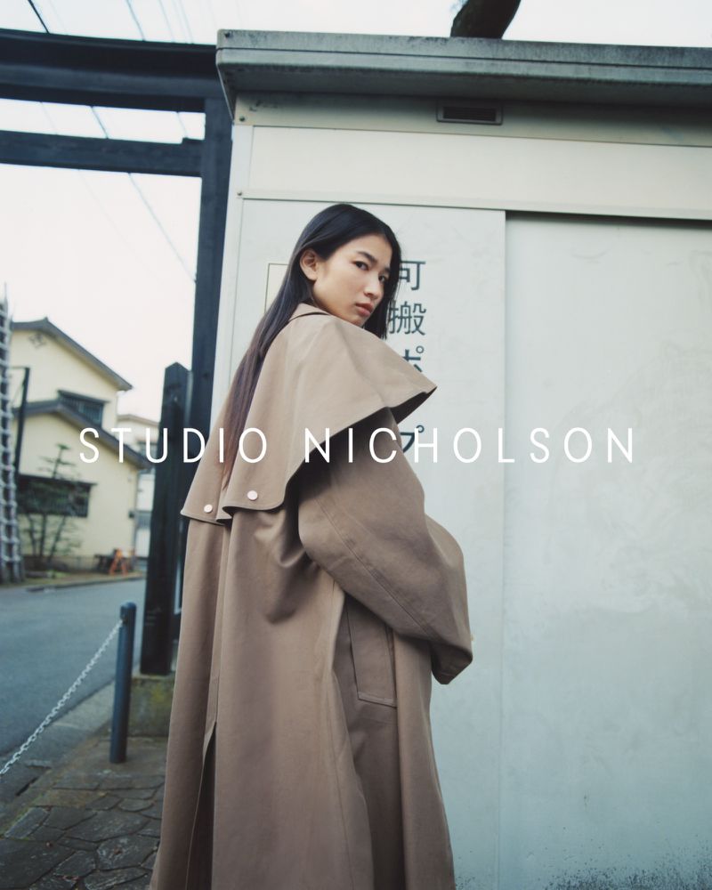Asian woman in a tan trench coat looking over her shoulder, posing in front of a white building and gray skies outdoors.