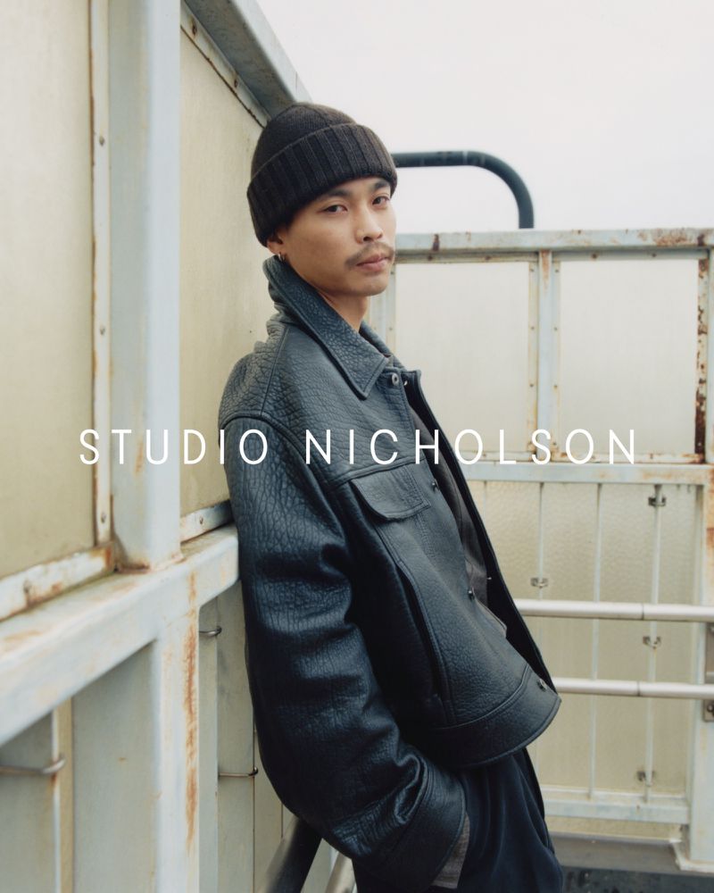 Portrait of an Asian male model in a black textured jacket and beanie leaning against a metal structure outdoors under overcast lighting.