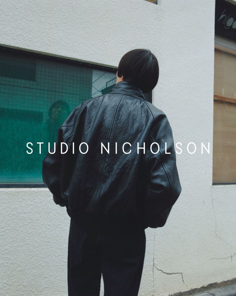 Back view of an adult wearing a black leather bomber jacket and black pants stands in an urban environment with white and tan walls.
