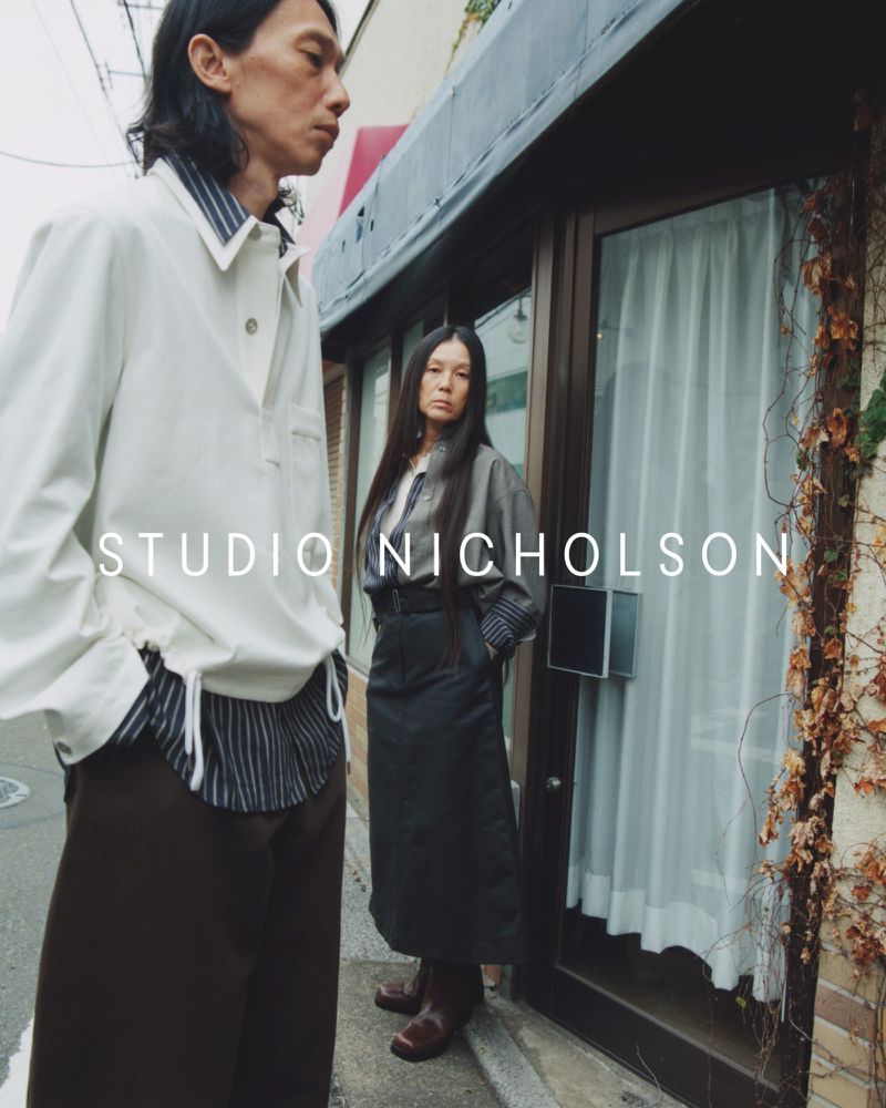 A man and a woman are standing in front of a building with dried vine. The man is in white, while the woman is in dark clothing.