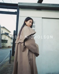 Model wearing an oversized taupe trench coat with a wide capelet collar looking back over her shoulder outdoors.
