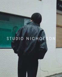 Rear view of a person with short dark hair wearing an oversized black leather jacket standing against a white textured exterior wall.