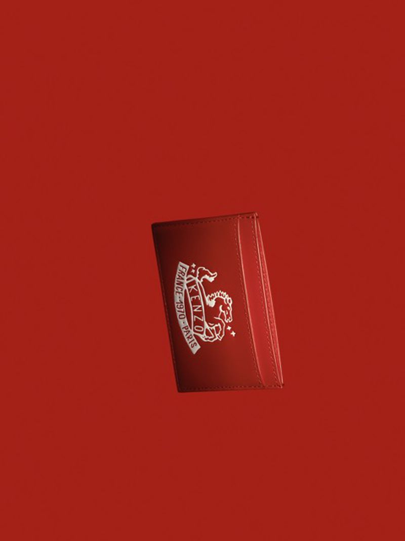 Close-up of a red leather card holder with a white logo floating against a solid red background, creating a minimalist studio product shot.